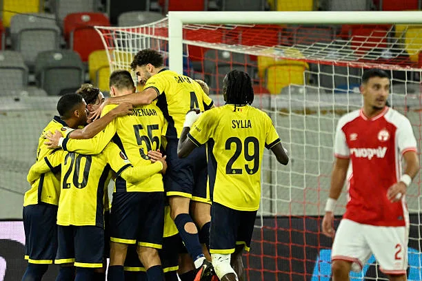 Ligue Europa - L’Union Saint-Gilloise déroule face à l’Hapoël Beer-Sheva (4-0) - wiwsport Union SG's Austrian forward #30 Raul Florucz (hidden) is celebrated by team mates after scoring by penalty the 1:0 goal as Hapoel Beer Sheva's Israeli defender #02 Guy Mizrahi (R) reacts during the UEFA Europa League league phase matchday 3 football match between Royale Union Saint-Gilloise and Hapoel Beer Sheva in Duesseldorf, western Germany, on October 22, 2026. (Photo by INA FASSBENDER / AFP via Getty Images)